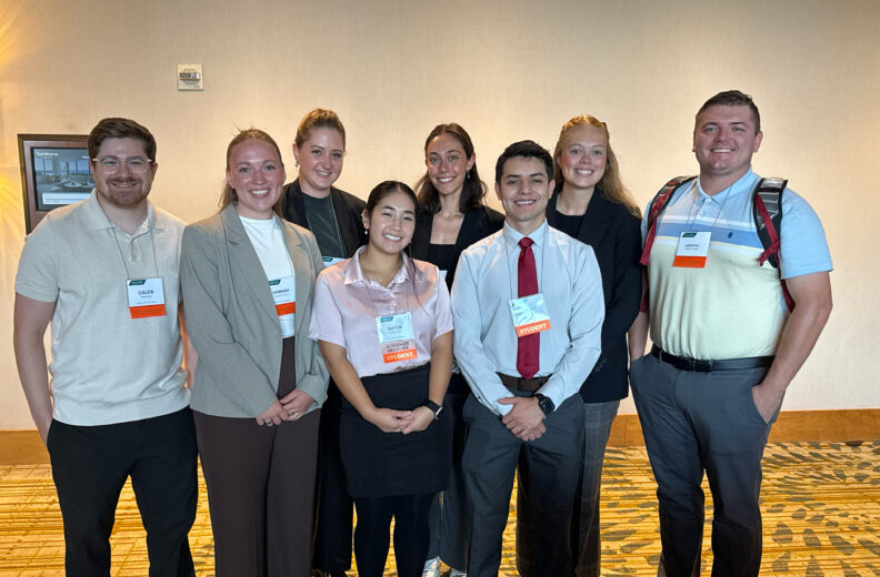 Eight individuals stand indoors, smiling at the camera. They are dressed in business casual attire and wearing name tags. The setting includes a beige wall, a framed picture on the left, and a patterned carpeted floor.