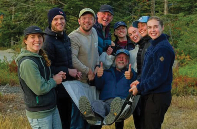 Group of eight people outdoors in a wooded area, smiling together. Two individuals hold up a person in the center on a makeshift seat, who is giving a thumbs-up. Everyone is dressed in outdoor clothing.