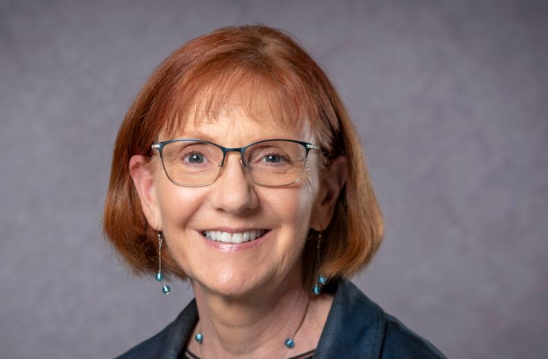 A person with short light brown hair and glasses smiles at the camera, wearing a dark jacket over a black top with turquoise earrings and a matching necklace. The background is a gradient from dark to light gray.