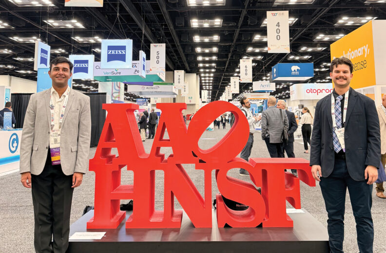 Two individuals stand on either side of a large red sculpture featuring the letters “AAO-HNSF” at a convention or trade show. The background shows a spacious exhibition hall with bright overhead lighting, multiple booths, and hanging signs displaying company names and booth numbers.