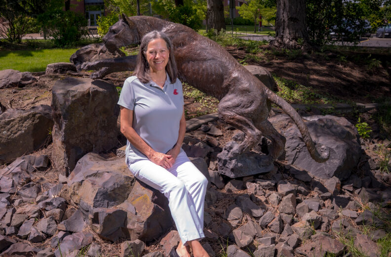 A person is seated on a cluster of large, uneven rocks in an outdoor setting with green grass and trees in the background. Behind the person is a bronze cougar statue in a dynamic pose, appearing to climb over the rocks. The person is wearing a light gray polo shirt with a red logo on the chest, white pants, and dark slip-on shoes. Sunlight casts strong shadows across the rocks and statue, highlighting their textures.