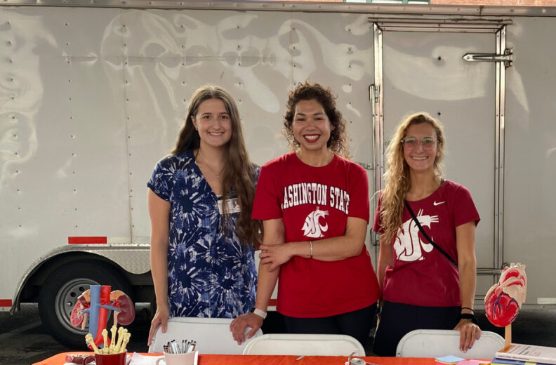 Three individuals stand behind a table covered with an orange tablecloth at an outdoor event. The table displays educational materials, including books, pamphlets, a stethoscope, anatomical heart models, and a mug with pens. Two people are wearing red Washington State University shirts with the WSU logo, and one person is wearing a blue patterned outfit. Behind them is a large silver trailer.