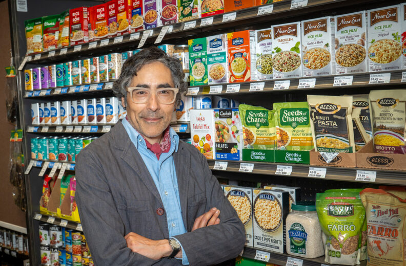A person stands with arms crossed in front of a grocery store shelf filled with various packaged grains, pasta, and seeds. The shelves display colorful boxes and bags labeled with brands such as Seeds of Change, Near East, and Lundberg. To the left, additional shelves hold snack products, including Tim’s chips. The setting appears to be inside a supermarket aisle with bright lighting.