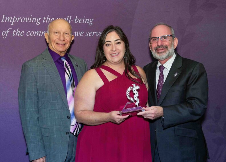 Three individuals standing together at an award ceremony in front of a purple backdrop with partial text reading ‘Improving the well-being of the comm… we serve.’ The person in the center is wearing a sleeveless burgundy dress and holding a clear glass award featuring a caduceus symbol. The individuals on either side are dressed in suits with patterned ties.