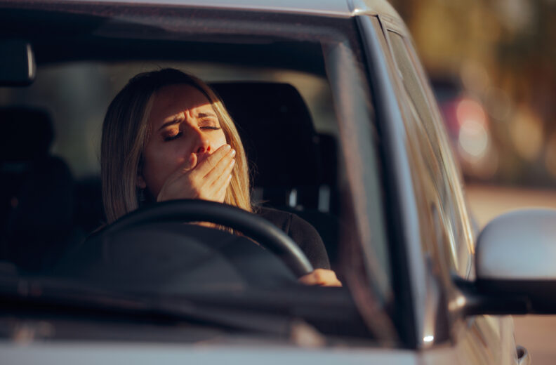 View from outside the front windshield of a parked vehicle, showing a person seated in the driver’s seat with hands on the steering wheel.