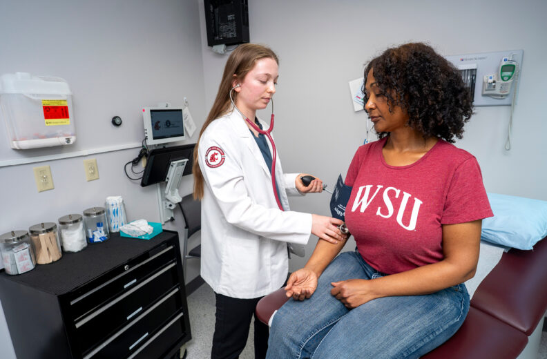 A healthcare professional wearing a white coat with a stethoscope around the neck is checking the blood pressure of a person seated on an examination table. The seated person is wearing a red WSU (Washington State University) T-shirt and jeans. The setting appears to be a medical examination room with equipment on the walls, a counter with medical supplies, and a blood pressure cuff on the patient’s arm.
