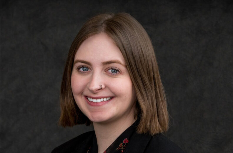 Professional headshot of a person with straight, shoulder-length light brown hair, wearing a dark blazer with a subtle floral pattern on the collar, set against a dark textured background.
