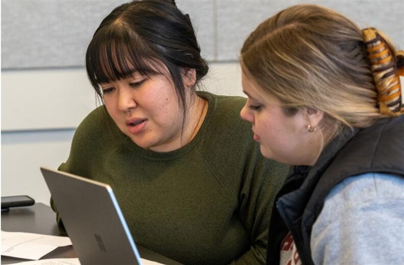 Two individuals seated at a table working together on laptops and papers. The setting appears to be a classroom or study area with gray walls.