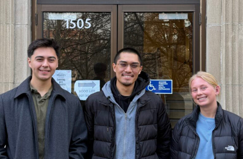 Three individuals standing in front of a glass door. The individuals are wearing winter jackets, and the building exterior features light-colored stone.