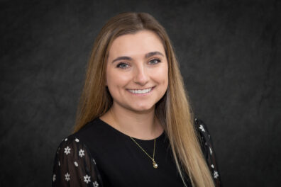 A professional portrait of a person with long, straight, light brown hair wearing a black top with sheer sleeves decorated with small white floral patterns. The individual is also wearing a delicate gold necklace with a small pendant. The background is a solid dark gray color.