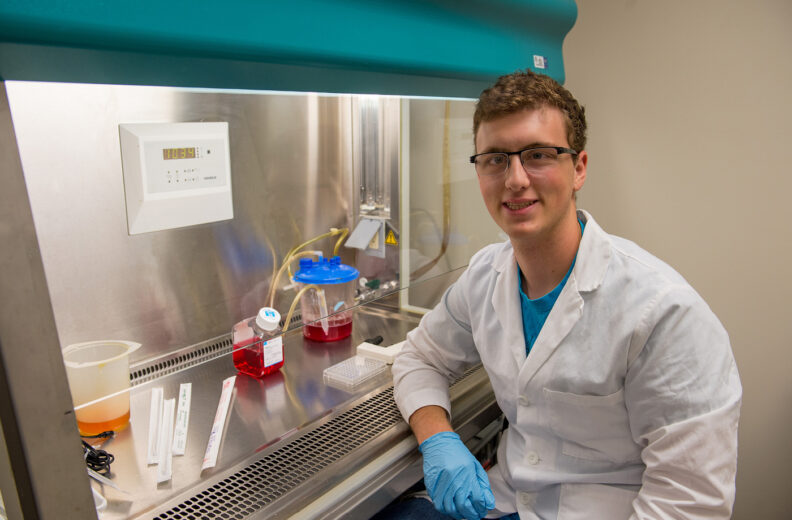 A person wearing a white lab coat and blue gloves is seated at a laboratory workstation. The workstation includes a biosafety cabinet with a teal-colored hood, two containers of red liquid, a clear container with tubing, a small tray, and several labeled strips. The background shows a beige wall and part of the cabinet’s control panel.