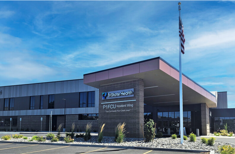 Exterior view of the TriState Health building under a clear blue sky. The entrance features a covered drop-off area with a sign reading “TriState Health P1FCU Inpatient Wing.” An American flag is mounted on a tall pole near the entrance, and landscaped plants line the walkway in front of the building.