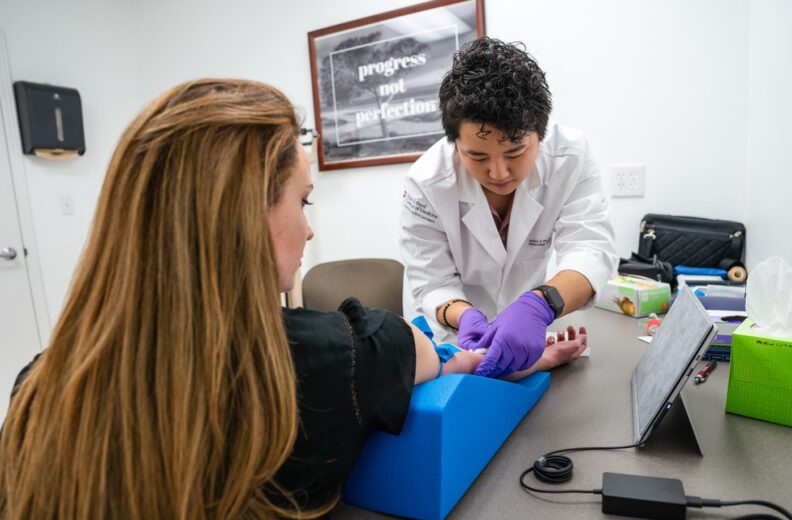 A healthcare professional wearing a white lab coat and purple gloves performs a procedure on a patient’s forearm, which is supported by a blue foam wedge on a desk. The desk holds a tablet, tissue box, and various medical supplies.