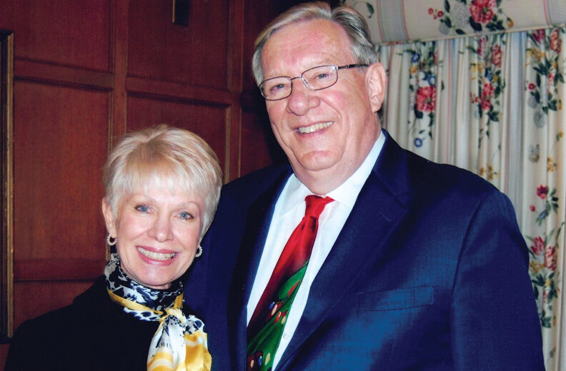 Two people standing close together in a formal indoor setting. One person is wearing a dark suit jacket with a white shirt and a festive tie featuring green and red colors. The other person is dressed in a dark outfit accessorized with a patterned scarf in shades of yellow, white, and black. The background includes wooden paneling and floral-patterned curtains.