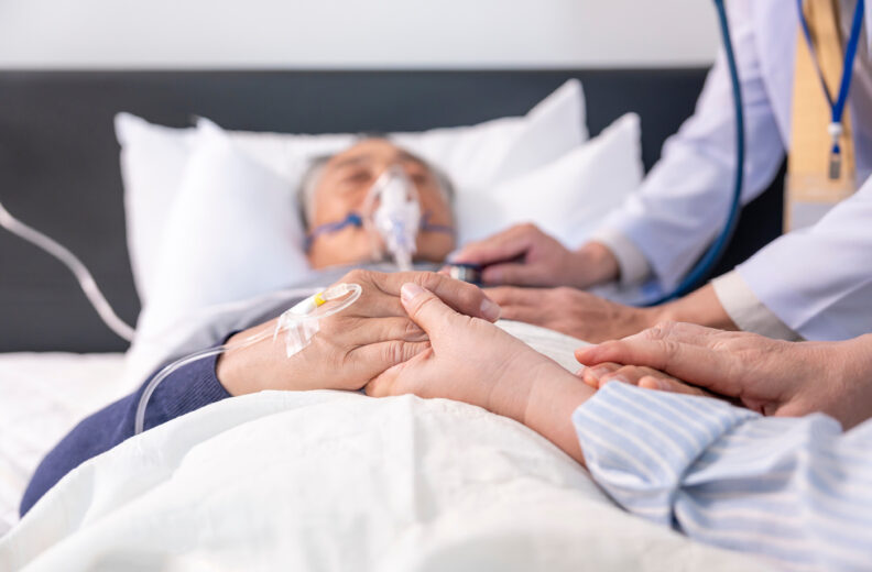 “A patient lies in a hospital bed with an IV in their hand while another person gently holds their hand. A healthcare professional stands nearby, offering support.