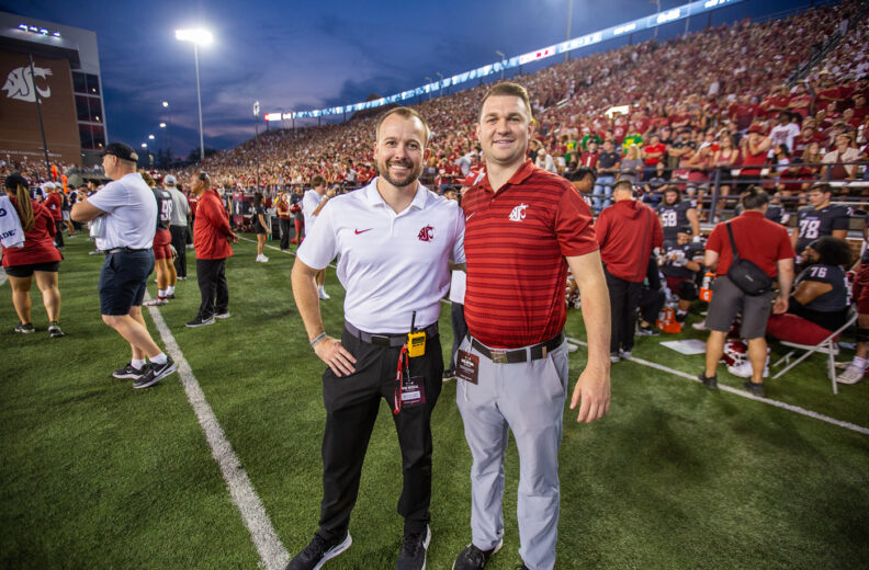 Two people wearing WSU-branded shirts stand on the sidelines of a football field during a game. A large crowd fills the stadium in the background as evening lights illuminate the scene.