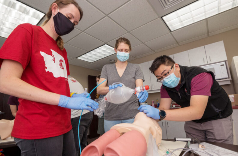 Three individuals wearing gloves practice a medical procedure on a training mannequin in a classroom setting. One person holds a ventilation bag, another manages a tube, and the third assists with positioning. The mannequin is placed on a table with visible anatomical features, and medical equipment is arranged nearby.
