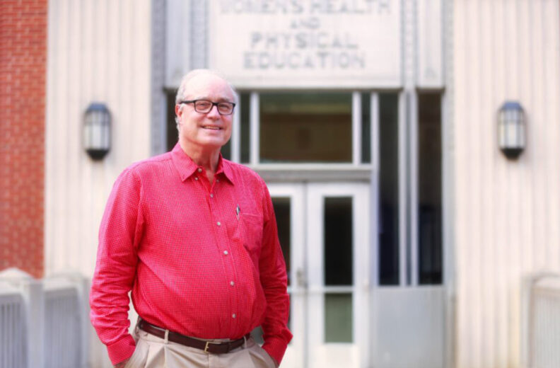 A person wearing a long-sleeved red checkered shirt and beige pants stands outdoors in front of a building entrance. The building has tall columns, brick walls, and a sign above the doors that reads “Health and Physical Education.” Two wall-mounted lamps flank the entrance, and the person has one hand in a pocket.