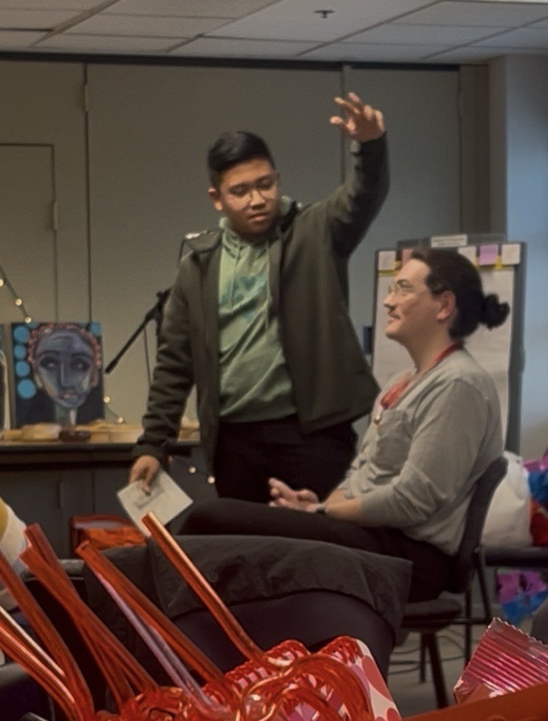 Two people are at the front of a room; one is standing and gesturing with an arm raised while holding a piece of paper, and the other is seated and facing forward.