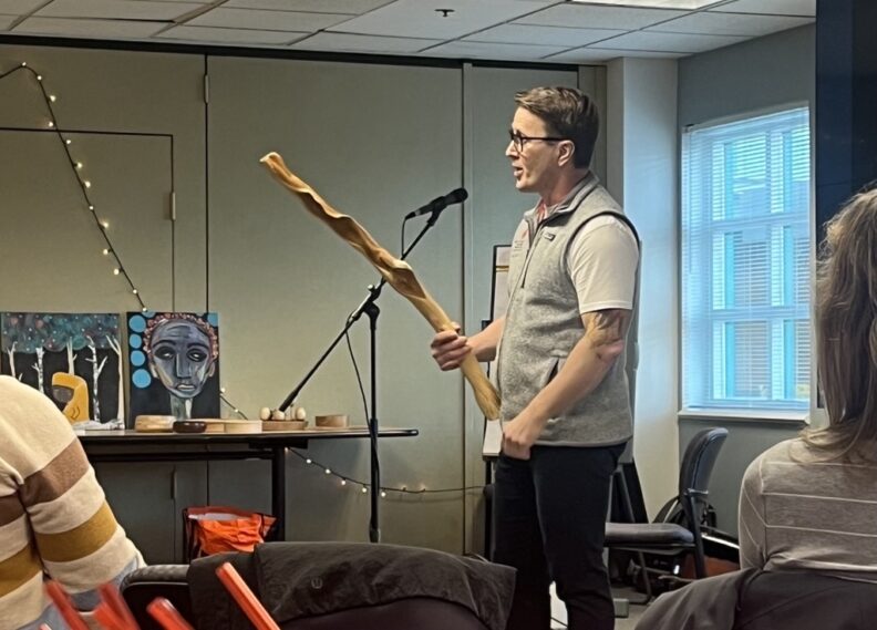 A person stands at the front of a room holding a large carved wooden staff while speaking into a microphone.