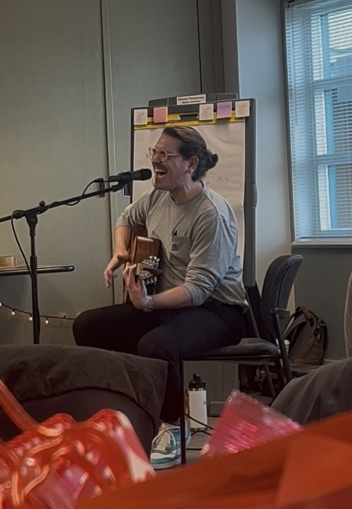 A person with glasses sitting down on a chair playing a guitar and singing.  