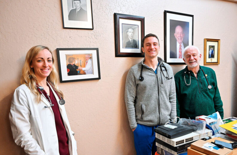 Three people wearing medical attire stand in an office space near a wall decorated with framed photographs and portraits. A cluttered desk with papers, equipment, and supplies sits in the foreground.