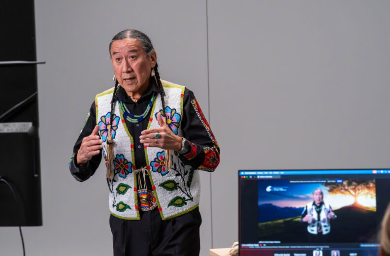 A person wearing a colorful beaded vest and traditional-style clothing is speaking or gesturing in a presentation setting. A nearby monitor displays a live video feed of the same person standing against a scenic virtual background.