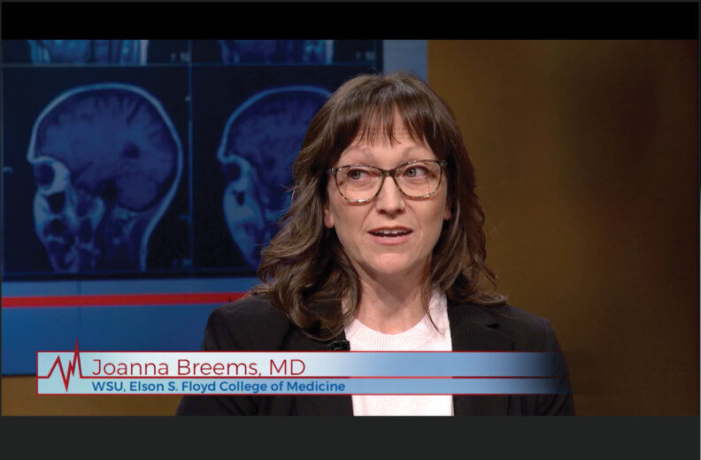 A person with shoulder‑length brown hair is shown speaking in a studio setting with medical brain-scan images displayed on a screen behind them. A lower-third graphic on the screen reads ‘Joanna Breems, MD – WSU, Elson S. Floyd College of Medicine.