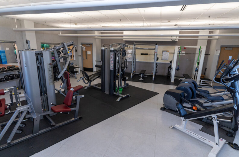 A spacious fitness room with various exercise equipment, including weight machines, free weights, cable machines, treadmills, and elliptical machines. The room has bright overhead lighting, light-colored flooring, and multiple doors along the back wall.