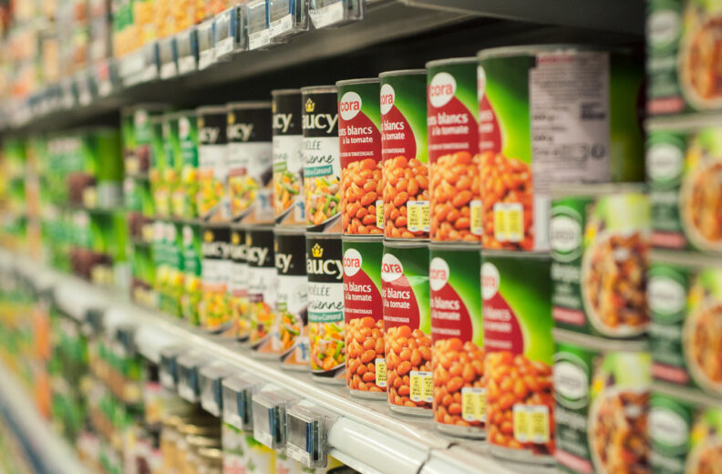 A grocery store shelf stocked with various canned foods, including beans and vegetables, arranged in rows with colorful labels.