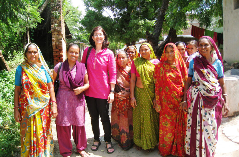 A group of people stand together outdoors on a sunny day, surrounded by trees and greenery. They are wearing brightly colored, patterned garments, including long draped fabrics and layered outfits in shades of yellow, red, green, purple, and blue. One person near the center is wearing a long-sleeved pink top and dark pants. The group is positioned on a dirt path with a small brick structure and foliage in the background.