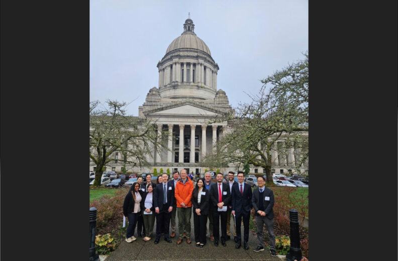 A group of people stands together for a photo in front of the Washington State Capitol building. The domed Capitol rises behind them on an overcast day, framed by leafless trees and parked cars along the drive.