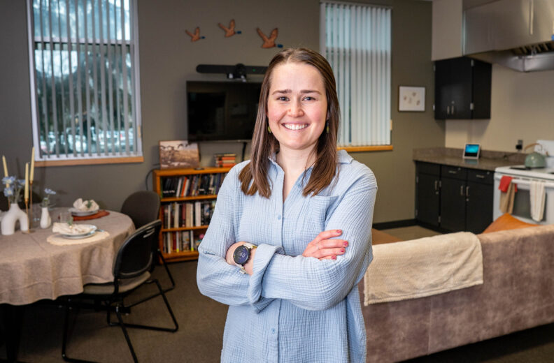 A person stands with arms crossed in a home‑like room that includes a small dining table, bookshelves, and a kitchen area. The person is wearing a long‑sleeved textured shirt and a watch. The background shows windows with blinds, wall décor, and various household items on the counters and shelves.