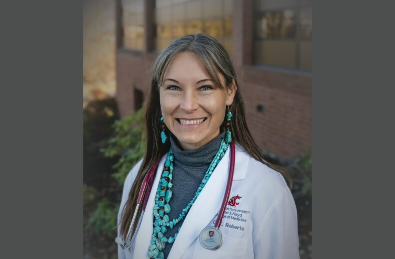 A healthcare professional wearing a white WSU College of Medicine coat and turquoise beaded necklaces stands outdoors in front of a brick building. A stethoscope hangs around their neck.