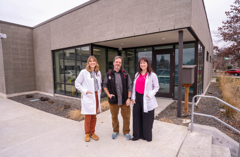A group of three people stands outside a modern clinic building with large windows and a concrete walkway. Two individuals wear white lab coats, and the third is dressed in casual professional clothing with a stethoscope around the neck. The building features gray brick walls, landscaped gravel areas, and an accessible ramp with metal railings.