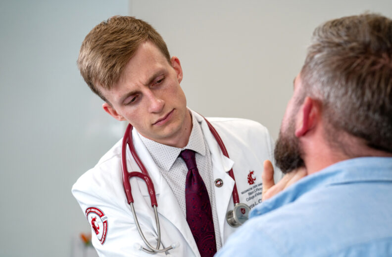 A medical student or clinician wearing a white coat with a stethoscope around the neck examines a patient’s throat or jaw area during a clinical assessment. The patient, seated in the foreground, is wearing a light blue shirt. The background is a plain clinical setting.