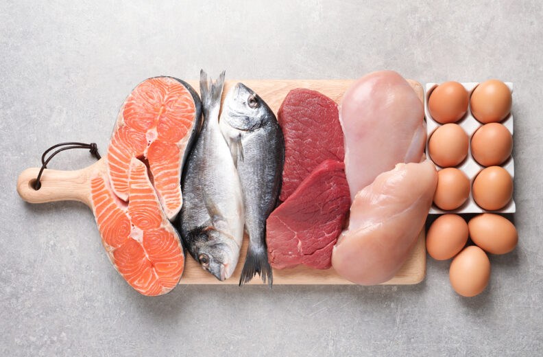 A wooden cutting board on a gray surface displays a variety of protein-rich foods, including salmon fillets, two whole fish, raw red meat, and raw chicken breasts. To the right of the board is a tray holding a dozen brown eggs.
