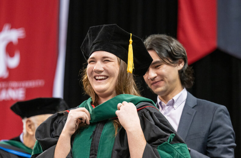 A graduation ceremony scene where an individual dressed in academic regalia is having a green hood placed over their shoulders by another person. Additional faculty in academic robes stand in the background, with large red and black banners hanging behind them.