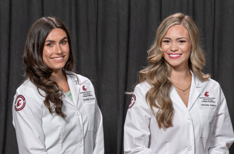 Two individuals wearing white medical-style lab coats stand in front of a dark curtain backdrop. Each coat displays embroidered text and logos associated with Washington State University’s College of Medicine. Both individuals have long, styled hair, and they are positioned side by side, facing the camera.