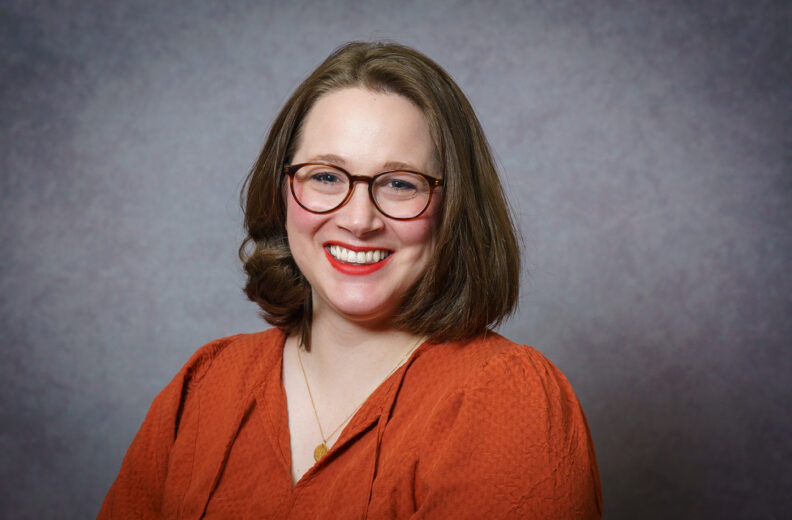 A person with shoulder‑length hair is pictured in a studio portrait setting against a gray textured backdrop, wearing an orange long‑sleeve blouse and a simple necklace.