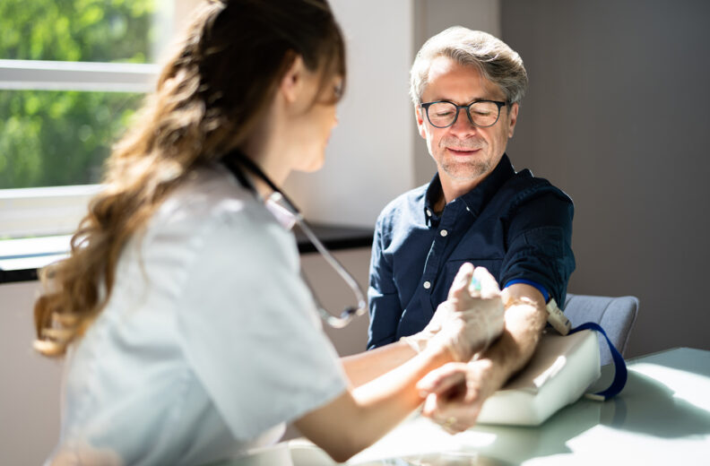 A healthcare professional measures an adult’s blood pressure using an arm cuff in a brightly lit exam room.