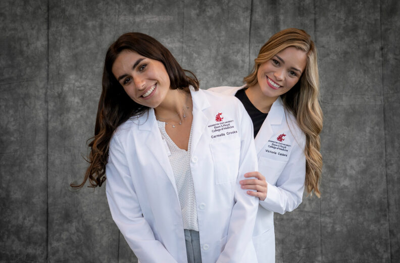 Two individuals wearing white lab coats stand together in front of a gray studio backdrop.