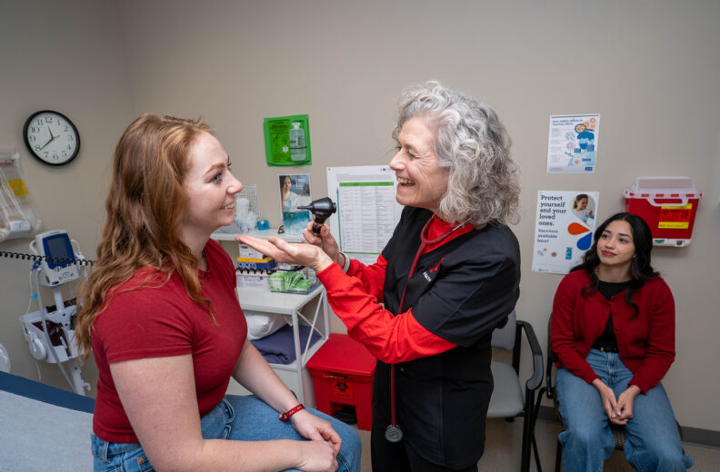 A healthcare provider examines a young woman’s eyes in a medical exam room while another student sits nearby observing.
