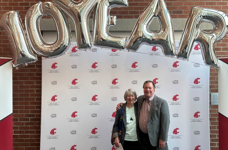 Two people stand in front of a step-and-repeat backdrop with Washington State University logos, beneath large silver balloons spelling “10 YEAR.”