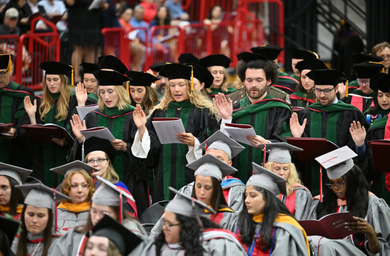 Graduates in caps and gowns raise their right hands and read from programs during a ceremony.