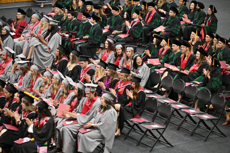Rows of graduates in caps and gowns seated and applauding indoors.