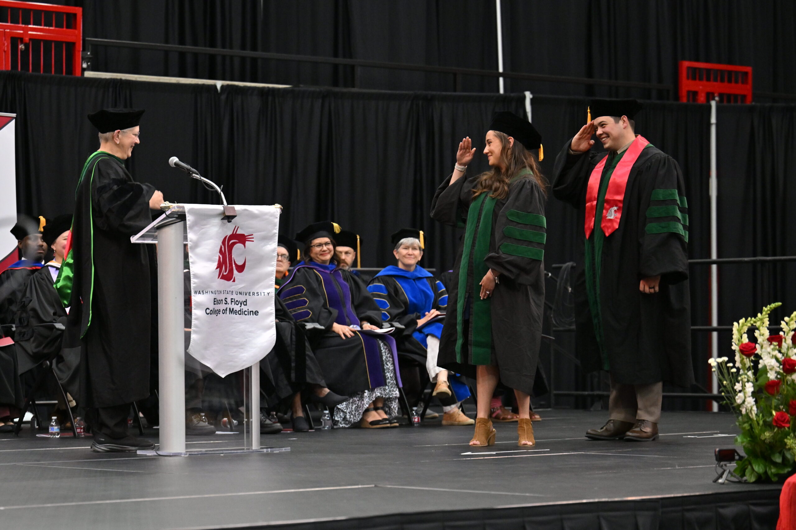 Two graduates saluting a professor sending behind a podium.