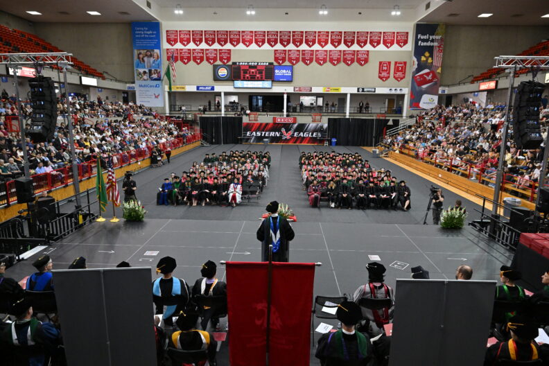 Wide view of a graduation ceremony in a crowded arena with a speaker at a podium.