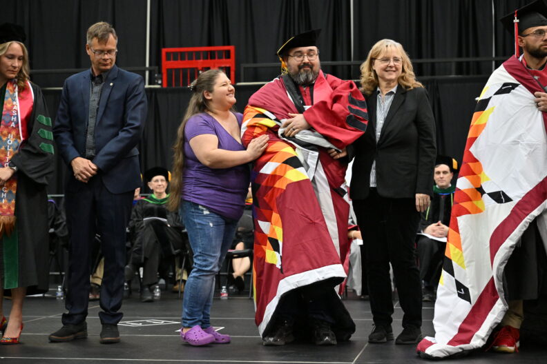 Graduate wrapped in a patterned stole stands on stage with others during a ceremony.