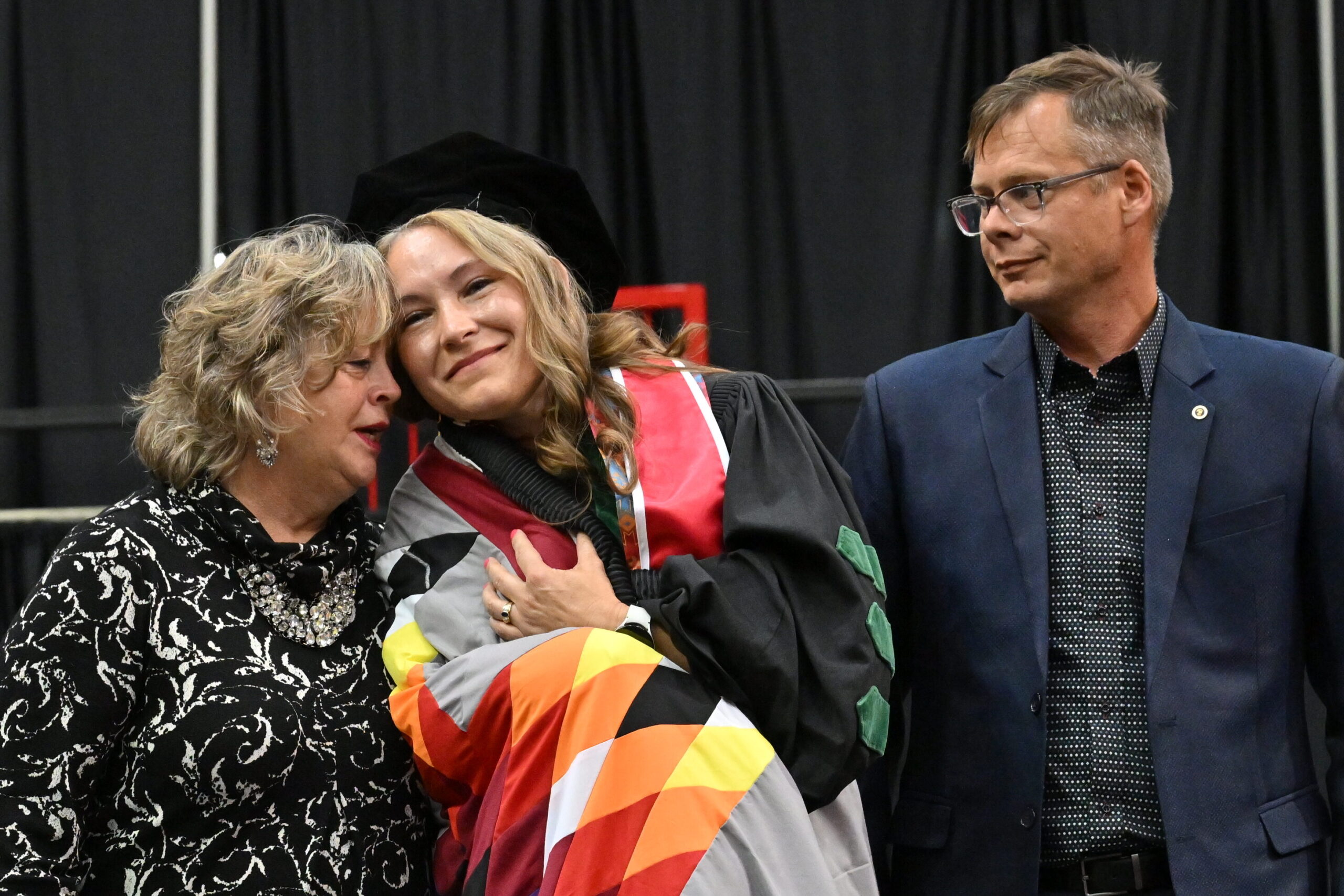 Graduate in cap and gown leans against another person on stage during graduation.